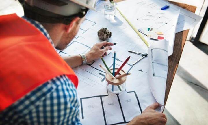 Een persoon met een helm en oranje vest bekijkt architectonische blauwdrukken op een tafel. Potloden in een houder, een rolmaat en andere planningsmaterialen liggen verspreid.