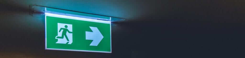 Verlicht groen nooduitgangbord met een wit pictogram van een persoon die door een deur loopt, vergezeld door een pijl naar rechts die de richting naar de uitgang aangeeft. Het bord is op een donker plafond gemonteerd.