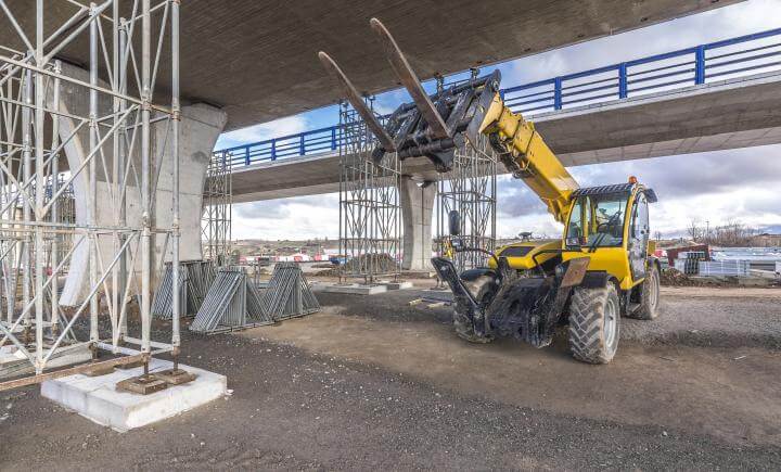 Een gele verreiker tilt veilig materialen onder een onafgemaakte brug. Steigers ondersteunen de constructie, omringd door bouwmachines en materialen, wat zorgt voor veilig werken. De lucht is gedeeltelijk bewolkt.