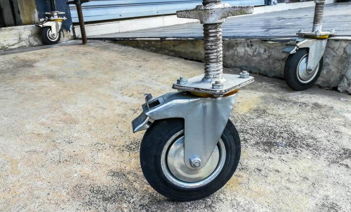 Close-up van drie industriële zwenkwielen op een betonnen oppervlak, bevestigd aan metalen bases met veren. De wielen, die doen denken aan die welke worden gebruikt in situaties zoals het monteren van een rolsteiger, lijken deel uit te maken van een grotere constructie, mogelijk voor zware apparatuur of meubels.