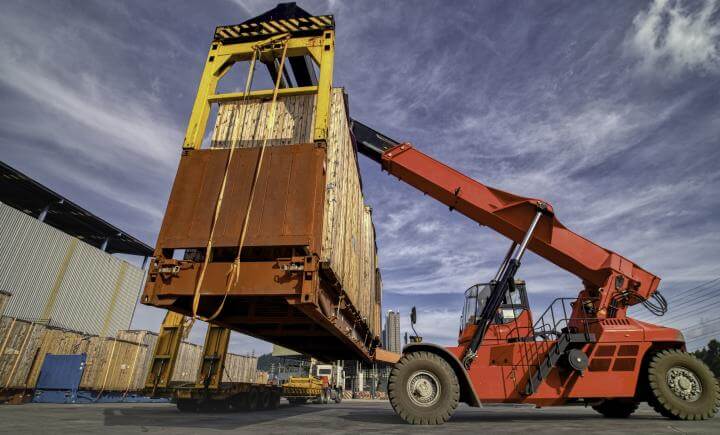 Een rode verreiker tilt veilig een grote, verroeste scheepscontainer op in een industriegebied. De lucht is gedeeltelijk bewolkt, met meer containers en gebouwen op de achtergrond.