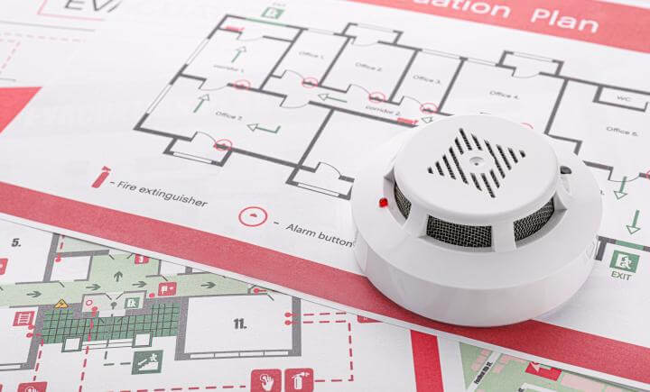 Een zelf maken rookmelder wordt op een evacuatieplan geplaatst, met pijlen voor vluchtroutes, brandblusserlocaties en alarmknoppen. Noodsymbolen en vluchtwegen zijn duidelijk gemarkeerd.