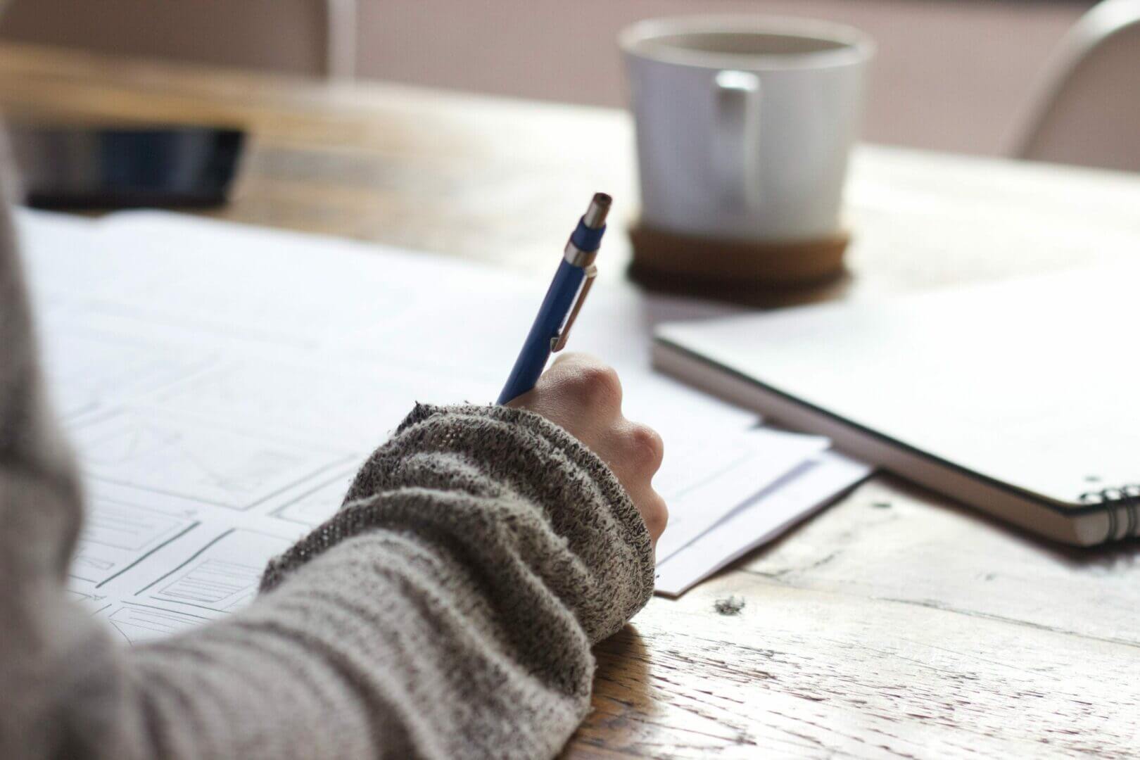 Een persoon in een grijze trui schrijft op papier met een blauwe pen. De scène bevat een witte mok op een onderzetter en een gesloten notitieboek op een houten tafel. De focus ligt op de hand en de pen, met een gezellige sfeer die wordt gesuggereerd door de kleding en de omgeving.