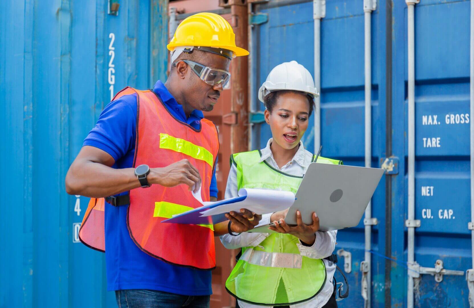 Twee werknemers met veiligheidshelmen en vesten staan bij scheepscontainers. De een houdt een klembord vast en de ander een laptop. Ze lijken iets te bespreken of te beoordelen dat verband houdt met hun werk.