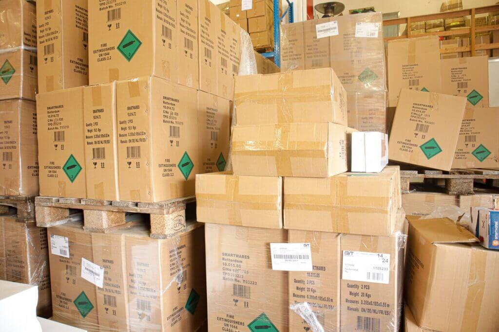Stapels ingepakte kartonnen dozen op houten pallets zijn te zien in een magazijn. De dozen zijn gelabeld met groene gevarensymbolen. De achtergrond toont planken gevuld met meer dozen en containers. Het gebied lijkt druk en georganiseerd.