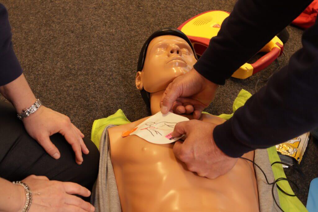 Twee personen demonstreren de AED-cursus, waarbij ze elektroden op de borst van een reanimatiepop aanbrengen, terwijl het AED-apparaat op de achtergrond zichtbaar is.