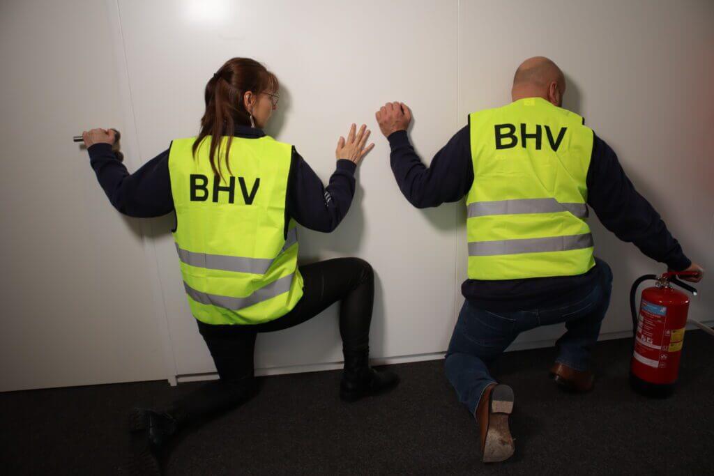 Twee personen met fluorescerende hesjes met het opschrift BHV knielen voor een witte muur, één met een brandblusser in de hand. Ze lijken een noodprocedure te simuleren alsof ze een zelf ontruimingsplattegrond maken.
