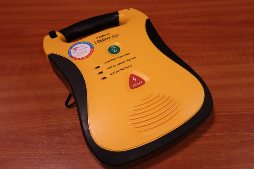 Een geel Lifeline AED (Automated External Defibrillator) apparaat staat op een houten oppervlak, met een handvat, knoppen en pictogrammen die verschillende functies aangeven, zoals het shock-symbool. Kennis van 6 levensreddende tips bij reanimeren kan het effectieve gebruik ervan tijdens noodsituaties verbeteren.