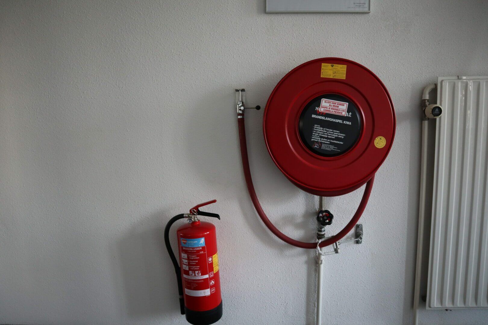 Een rode brandslanghaspel gemonteerd op een witte muur met daaronder een brandblusser benadrukt de veiligheidsvoorbereiding. Aan de rechterkant is een radiator zichtbaar, wat drie redenen onderstreept om een beheerder brandmeldinstallatie cursus te volgen: uitgebreide kennis, naleving en efficiënte respons.