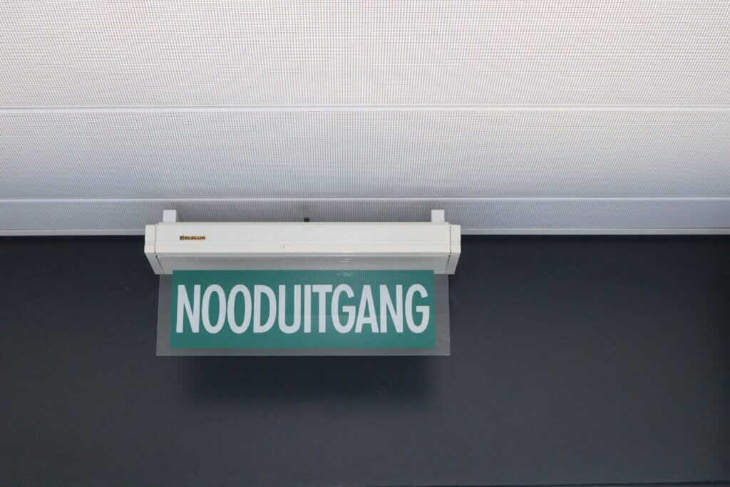 Een groen bord met de witte tekst NOODUITGANG, wat in het Nederlands NOODUITGANG betekent, is gemonteerd op een donkere muur onder een wit plafond. Dit is wat een ontruimingsoefening kan voorkomen.