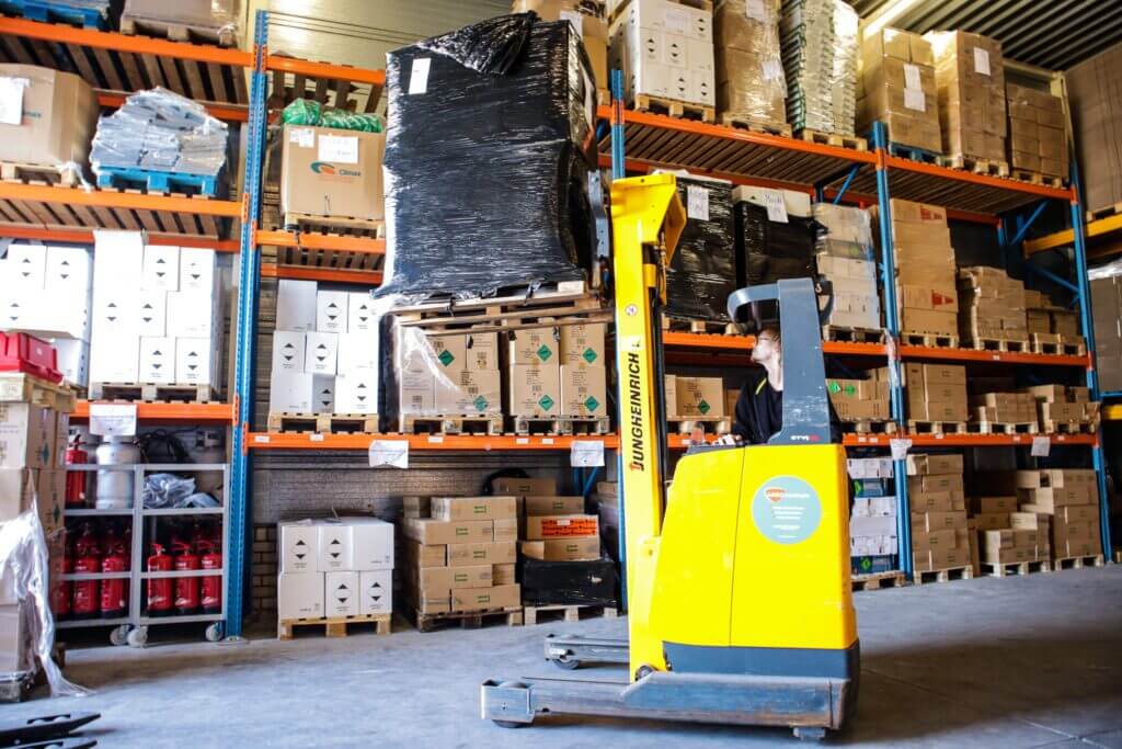 Een felgele heftruck tilt een pallet ingepakt in zwart plastic naar een hoge plank in het drukke magazijn. Terwijl reachtruckchauffeurs aan de slag gaan, staan er verschillende dozen op de planken en worden brandblussers netjes opgestapeld in de hoek.