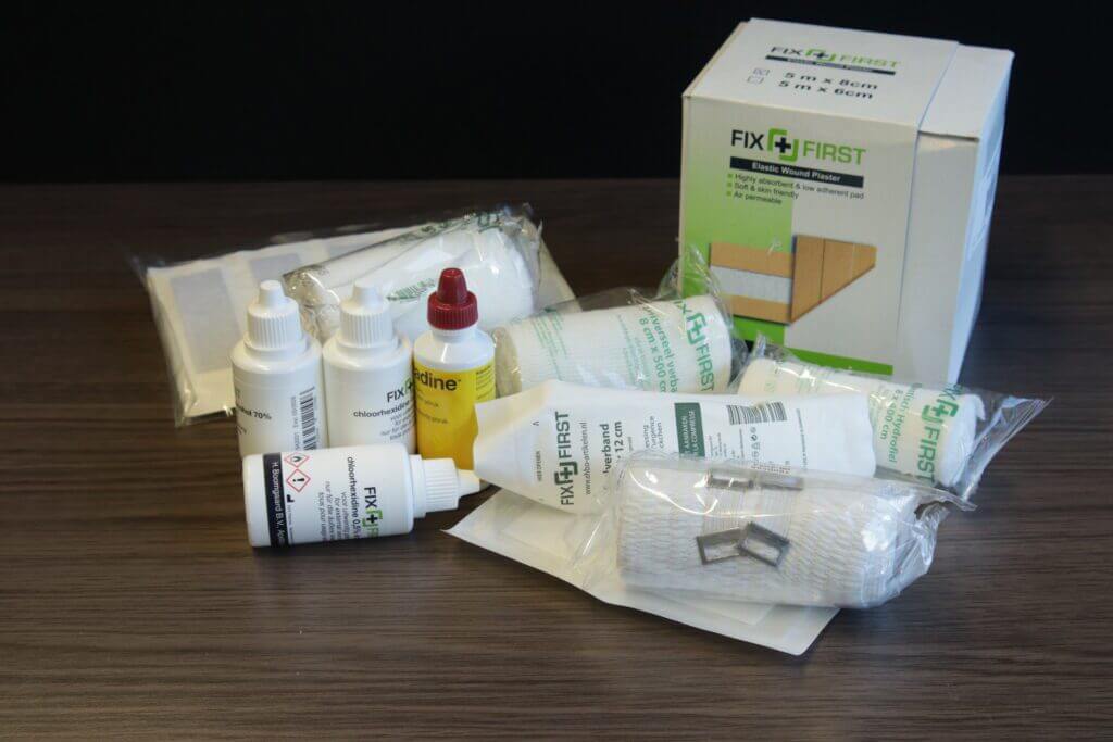 Een verzameling eerstehulpmaterialen staat op een tafel, met verbanden, flessen met antiseptische oplossingen en gaasverbanden. De FIX FIRST-merkdoos bevat essentiële benodigdheden en tips voor het goed verzorgen van een open wond, zodat u voorbereid bent op kleine noodgevallen.