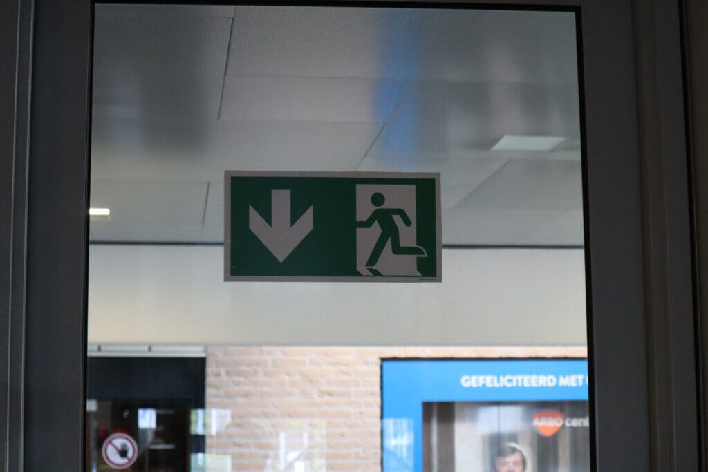 Een glazen deur toont een groen en wit nooduitgangbord, met een pijl naar beneden en een figuur die naar een deur rent. In het wazige interieur met een bakstenen muur en poster lijkt het op een ontruimingsoefening voorbeeld.
