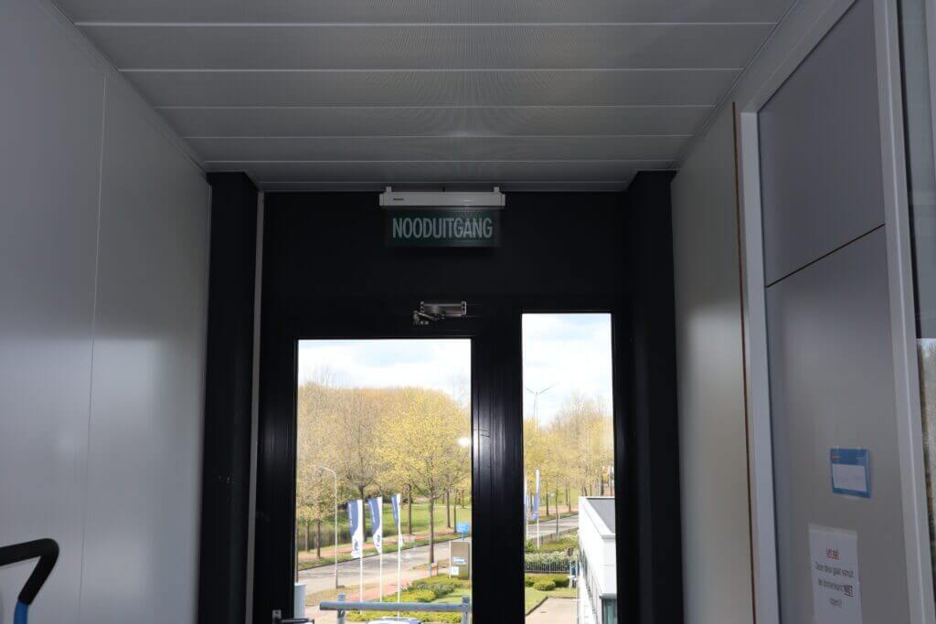 Een glazen deur met het opschrift NOODUITGANG (wat nooduitgang betekent in het Nederlands) leidt naar buiten, wat zorgt voor een snelle en veilige evacuatie. Het uitzicht toont een pad met bomen en vlaggenmasten onder een bewolkte lucht, allemaal badend in helder natuurlijk licht.