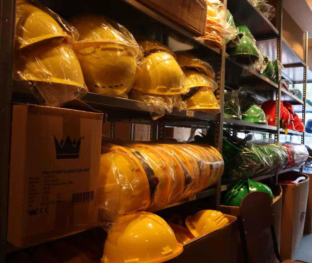 Planken met stapels gele en groene bouwhelmen in plastic verpakt, naast kartonnen dozen. De kamer is natuurlijk verlicht en toont verschillende veiligheidsuitrustingen voor de bouw. Dit zijn de bedoeling van de kleuren bouwhelmen omdat ze verschillende rollen op een bouwplaats symboliseren.