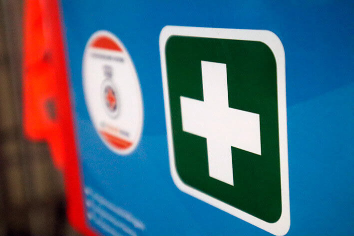 Close-up van een blauw bord met een wit kruis op een groene achtergrond, dat symbool staat voor eerstehulptips zoals Hoe brandwonden behandelen? Links staat een cirkelvormig embleem met rode en witte details, maar het is gedeeltelijk onscherp.