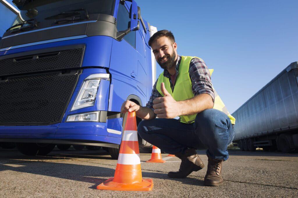 Een man in een geel veiligheidsvest hurkt en steekt zijn duim omhoog naast een oranje verkeerskegel voor een blauwe vrachtwagen. Zijn glimlach suggereert tevredenheid of goedkeuring, wat de geest van dit is waar code 95 voor dient belichaamt. De achtergrond toont meer vrachtwagens en een heldere lucht.