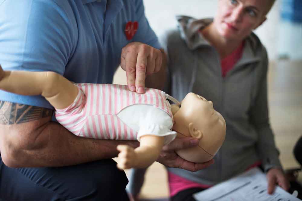Een persoon demonstreert reanimatie van een baby op een babypop, na een stappenplan reanimeren baby. Een andere persoon kijkt aandachtig toe en houdt een vel papier vast. De setting lijkt een reanimatietrainingssessie te zijn.