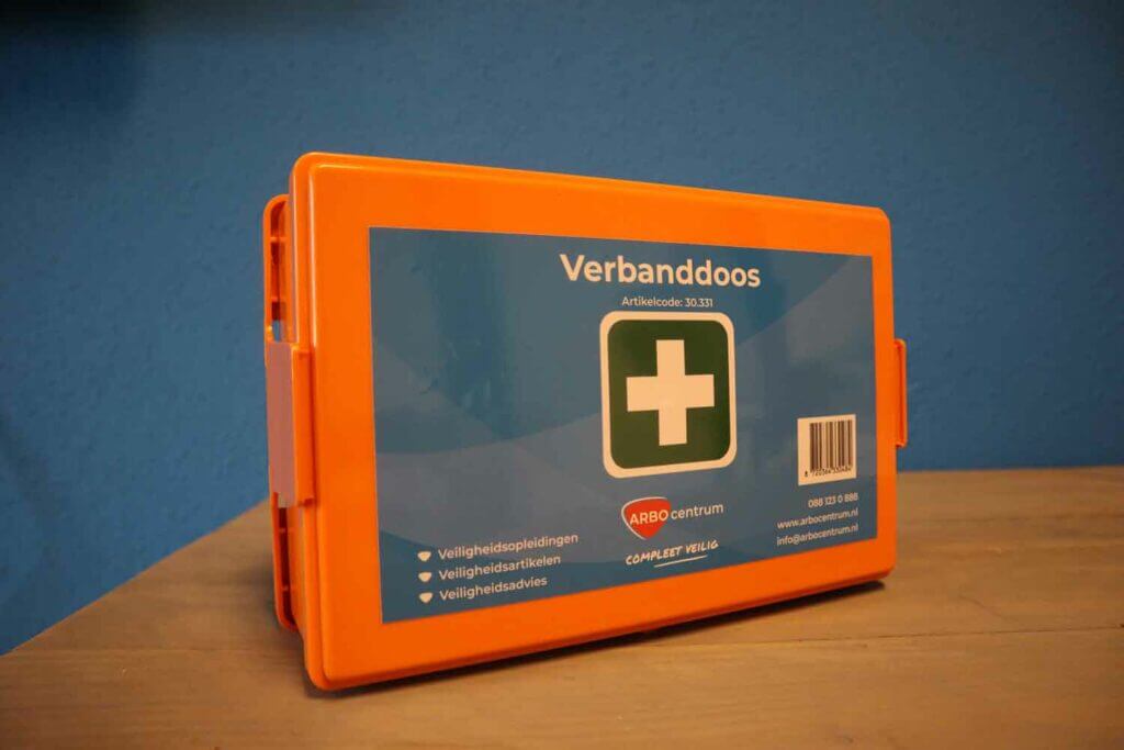 Een oranje EHBO-doos met het label Verbanddoos met een wit kruis op een groene cirkel rust op een houten oppervlak tegen een blauwe muur. Naast de essentiële noodvoorraden staat er 5 Redenen om een verbanddoos thuis te hebben en bevat het contactgegevens voor extra gemoedsrust.