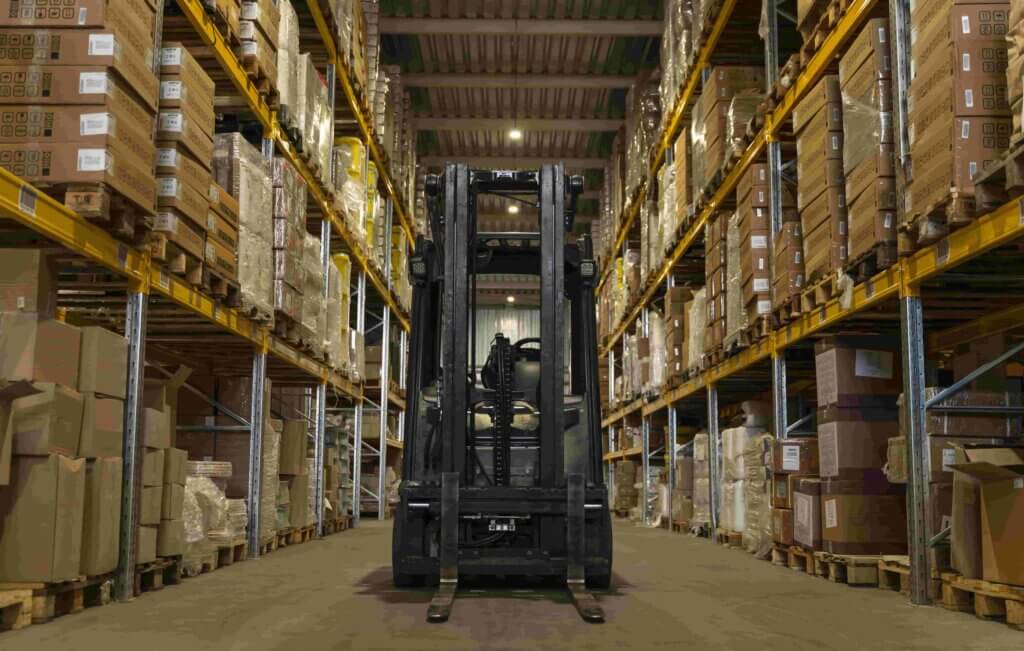 Een vorkheftruck staat geparkeerd in het midden van een magazijngang, omringd door hoge planken vol met verschillende dozen en pakketten. De planken zijn netjes georganiseerd, wat de voordelen van on-site vorkheftrucktraining laat zien, aangezien de verlichting in het magazijn een gerichte sfeer creëert.