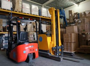 Twee vorkheftrucks, een rode en een gele, staan geparkeerd in een magazijn vol met stapels kartonnen dozen en pallets. Planken staan vol met grote ingepakte pakketten onder metalen balken en lampen, klaar voor de De heftruck en reachtruck cursus op 1 dag om de operationele efficiëntie te maximaliseren.