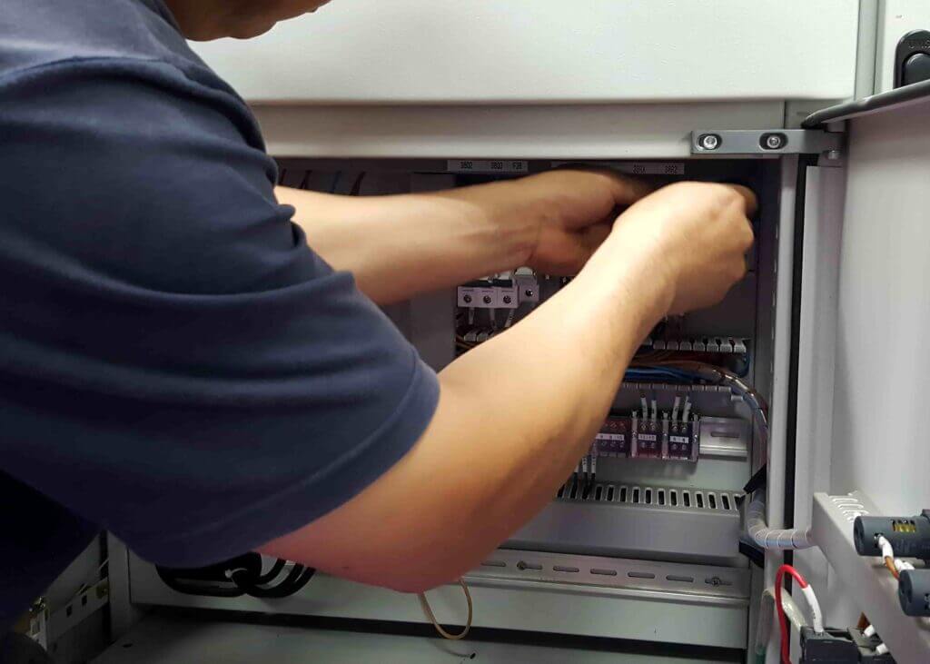 Een persoon in een donker shirt past vakkundig draden aan in een elektrisch paneel, omgeven door schakelaars en circuitcomponenten. Meesterschap als dit komt vaak voort uit het begrijpen van de voordelen van het NEN 3140 certificaat, dat veiligheid en naleving waarborgt.