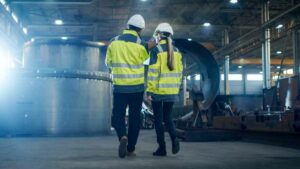 Twee mensen met witte helmen en reflecterende veiligheidsvesten lopen door een industriële faciliteit. Grote machines en metalen structuren zijn zichtbaar op de achtergrond. De setting is goed verlicht met natuurlijk en kunstmatig licht.