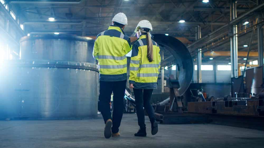 Twee mensen met witte helmen en reflecterende veiligheidsvesten lopen door een industriële faciliteit. Grote machines en metalen structuren zijn zichtbaar op de achtergrond. De setting is goed verlicht met natuurlijk en kunstmatig licht.