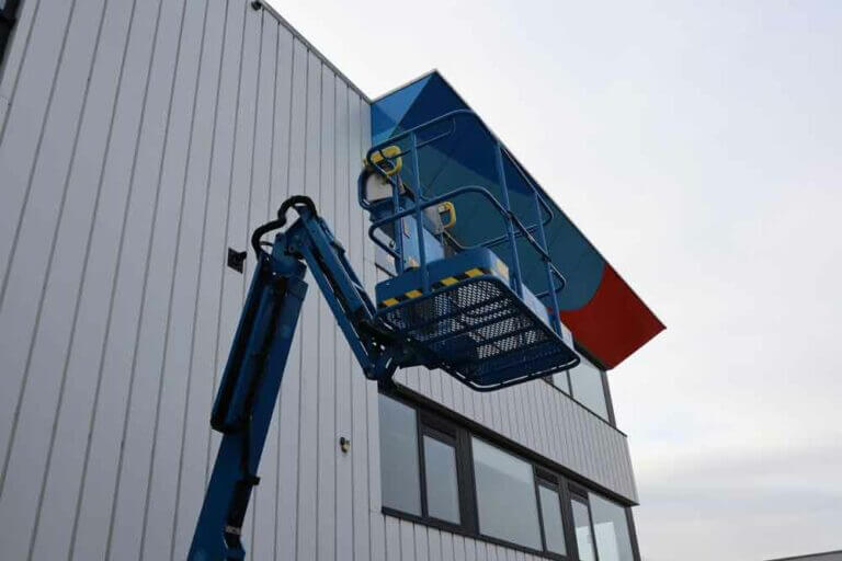 Een blauwe hoogwerkerlift staat naast een gebouw met witte verticale gevelbekleding en donkere ramen. Het platform van de lift bevindt zich bij een kleurrijke luifel boven de ramen, met blauwe en rode accenten. De lucht is bewolkt, wat illustreert waarom het behalen van een hoogwerkercertificaat essentieel is.