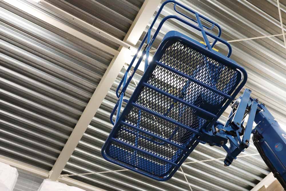 Tussen de 3 manieren om veilig op hoogte te werken, wordt een blauw schaarliftplatform omhoog gebracht naar een metalen plafond, het gaasoppervlak is duidelijk zichtbaar. De lange metalen panelen van het plafond worden ondersteund door beige balken, met verlichting die weerkaatst om heldere verlichting te bieden.