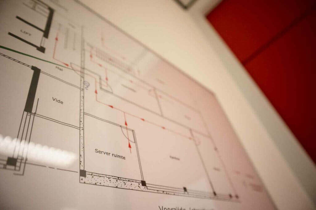 Een close-up van een technisch plattegrond voor een ontruimingsplan, bevestigd aan een muur. Het plan bevat gelabelde secties en kamers, met richtingspijlen en gemarkeerde metingen. Op de achtergrond zorgt de rode muur voor contrast, wat drie cruciale voordelen van een ontruimingsplan benadrukt.