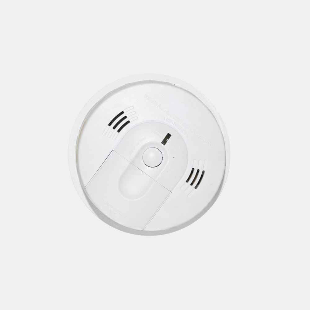 Een witte rookmelder met een ronde vorm, met een centrale knop en ventilatieopeningen aan beide kanten, is gemonteerd op een eenvoudig wit plafond. Het eenvoudige ontwerp benadrukt de functionaliteit en de veiligheidsvoordelen van het hebben van een beheerder voor uw brandalarmsysteem in een residentiële omgeving.
