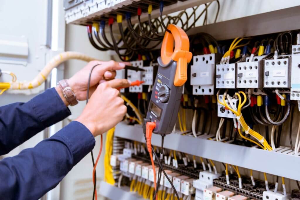De handen van een elektricien gebruiken vakkundig een multimeter met probes om elektrische verbindingen in een circuit breaker paneel te testen, wat het belang van het behalen van het NEN 3140 keurmeester diploma benadrukt. Het paneel is gevuld met draden en componenten, en de multimeter heeft een oranje klem.