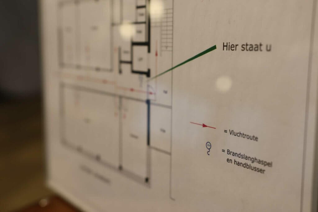 Close-up van een ontruimingsplan van een gebouw op een muur, met details over Hier staat u naast de vluchtroute en brandblussymbolen. Tekst is in het Nederlands, met nadruk op de 3 voordelen van een ontruimingsplan voor veiligheid en efficiënte ontruiming.