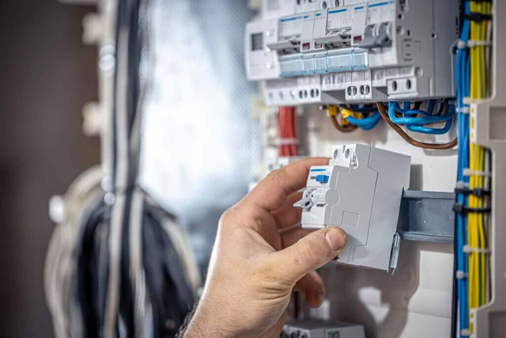 Een hand past een stroomonderbreker aan in een elektrisch paneel, wat illustreert waarom het essentieel is om een gecertificeerde NEN 3140-inspectie-expert te worden. De georganiseerde, kleurgecodeerde draden en componenten van het paneel vallen op tegen de wazige achtergrond, wat het belang van vakkundige bediening benadrukt.