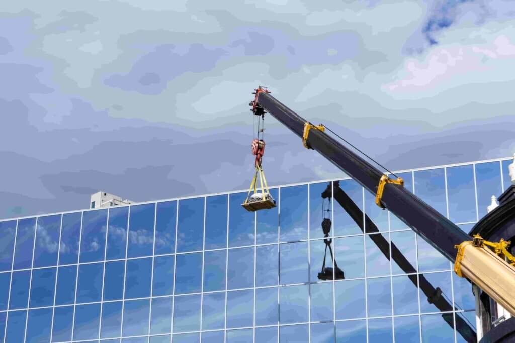 Een kraan tilt bouwmaterialen op voor een modern glazen gebouw met een bewolkte lucht op de achtergrond. De reflecterende ramen van het gebouw laten een gespiegeld beeld van de kraan zien.