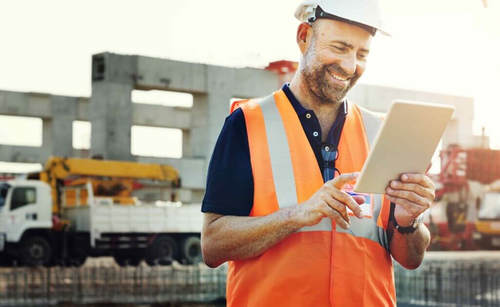 Een bouwvakker in een oranje veiligheidsvest en helm, zich bewust van de nadelige gevolgen als je geen VCA-diploma hebt, glimlacht terwijl hij een tablet gebruikt. Op de achtergrond rijst een gedeeltelijk gebouwde constructie op te midden van drukke bouwvoertuigen.