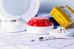 Verschillende brandveiligheidsvoorzieningen, waaronder rookmelders, een geel handbrandmelder en een ronde luidspreker, worden weergegeven op een set bouwtekeningen. Een witte USB-kabel is ook zichtbaar op de voorgrond.