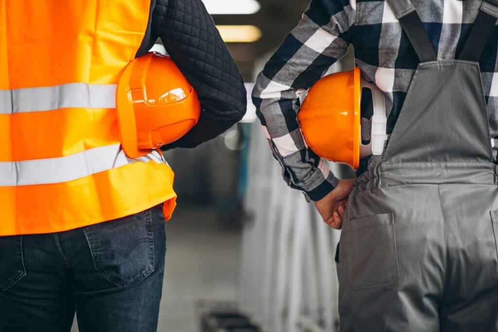 Twee bouwvakkers van achteren gezien, met oranje veiligheidshelmen in hun handen. De een draagt een oranje veiligheidsvest en de ander een geruit overhemd met grijze overall. Ze staan in een industriële omgeving, misschien een scène om over na te denken terwijl ze de 4 tips voor het slagen voor je VCA-examen overwegen.