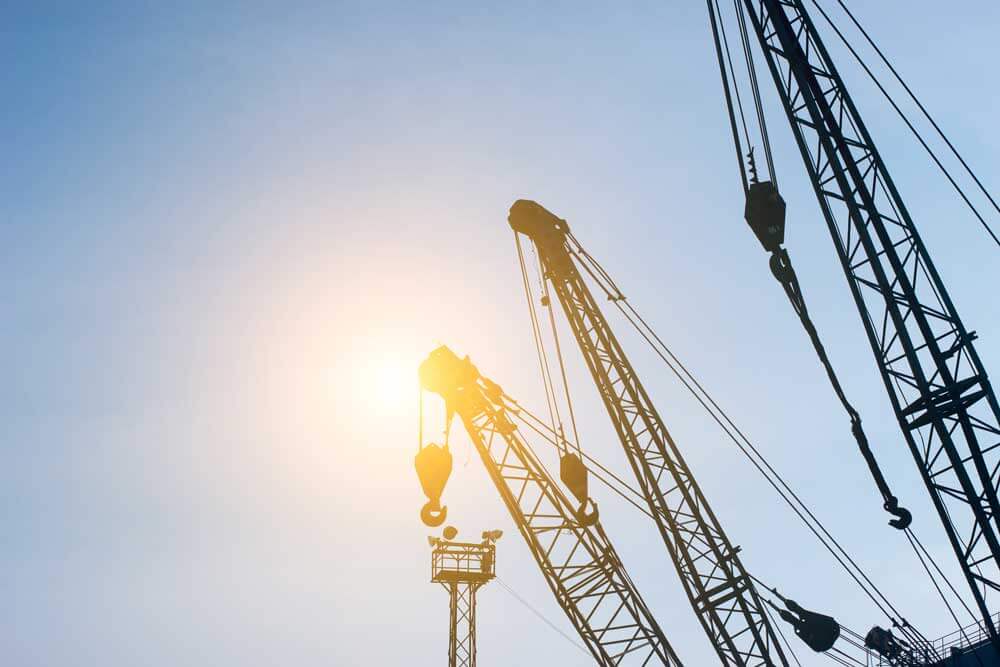 Silhouetten van bouwkranen tegen een strakblauwe lucht, met de ondergaande zon erachter die een warme gloed werpt. De kranen zijn hoog en hoekig, wat het nut van een veilig hijsen-certificaat op deze drukke bouwplaats of industrieterrein onderstreept.