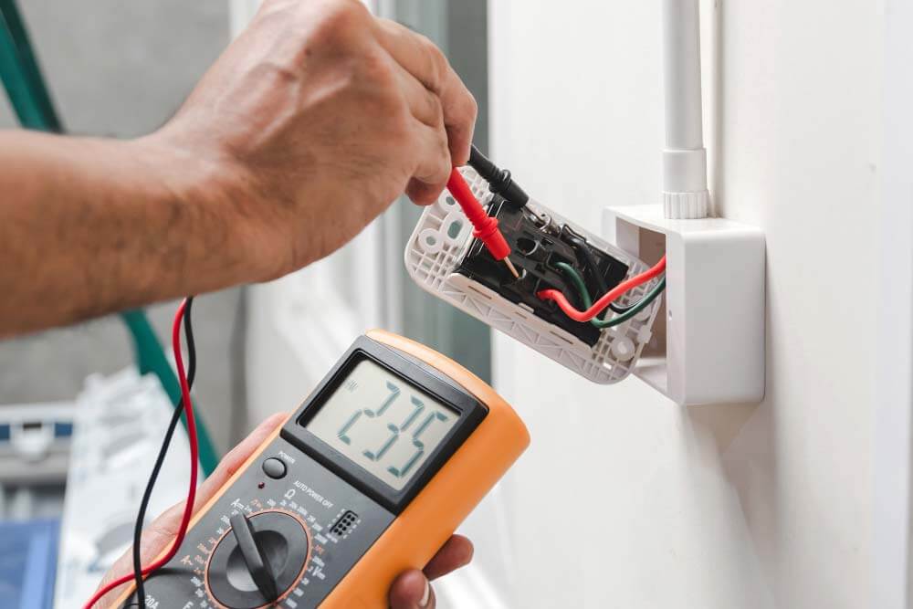 Een elektricien, die de voordelen van een NEN3140 keurmeester begrijpt, gebruikt een digitale multimeter om de spanning in een stopcontact te meten. Het scherm geeft 235 volt aan terwijl hij de probes vakkundig tegen de uitlopende draden houdt.