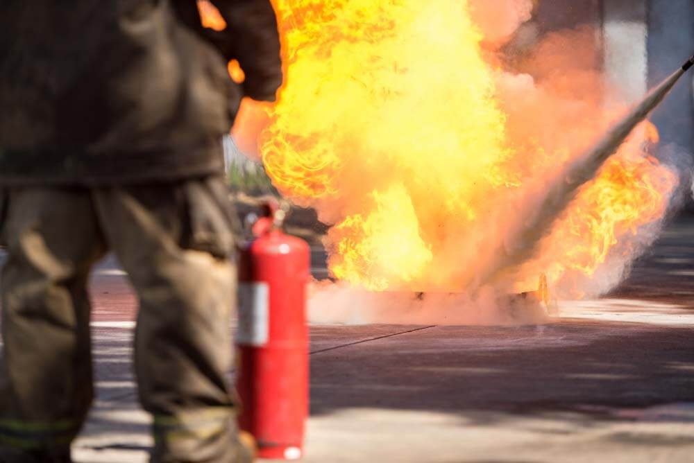 Een brandweerman, gedeeltelijk zichtbaar van achteren, houdt een rode brandblusser vast terwijl een grote vlam voor hem brandt. Een andere blusslang spuit het vuur, waardoor er een witte mist in het zonlicht op het betonnen oppervlak ontstaat. Dit laat zien waarom een beheerder van brandmeldinstallaties cruciaal is voor de veiligheid.