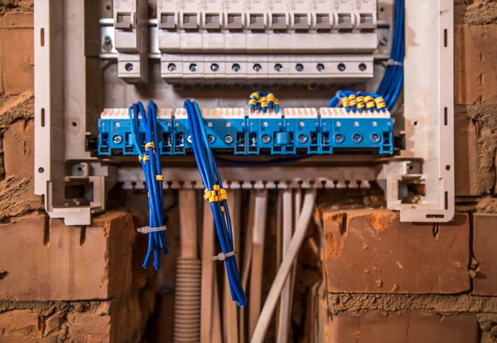 Elektrisch paneel met meerdere blauwe draden die zijn aangesloten op verschillende schakelaars, geplaatst in een bakstenen muur. De draden zijn gebundeld en gelabeld met gele labels. Volgens het principe Zo werk je veilig aan elektrotechnische installaties, lijkt het paneel open, met een nette en georganiseerde lay-out.