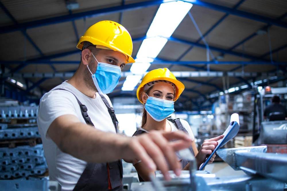 Twee arbeiders, beiden met gele helmen en blauwe gezichtsmaskers, staan in een fabriek. De man links wijst naar machines terwijl de vrouw rechts een klembord vasthoudt, en bespreekt hoe een preventiemedewerker de veiligheid en efficiëntie in deze industriële omgeving kan verbeteren.