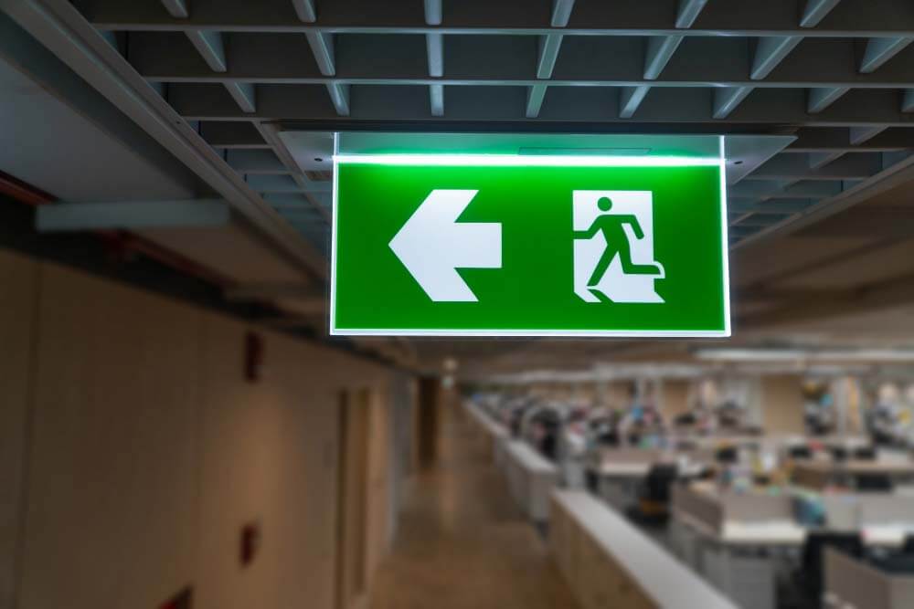 Een groen nooduitgangbord met een witte pijl die naar links wijst en een icoon van een rennende figuur is aan het plafond van een kantoorgang bevestigd, wat een van de 4 redenen om een ontruimingsplan te hebben benadrukt. De achtergrond toont wazige kantoorwerkplekken met bureaus en scheidingswanden.