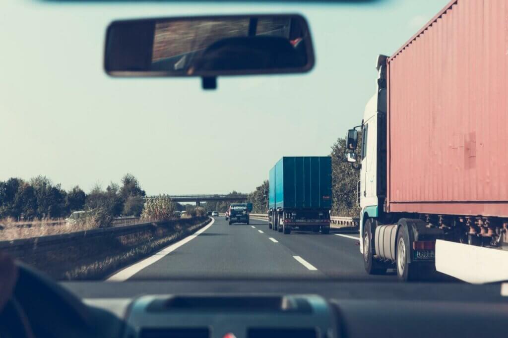 Vanuit het interieur van een auto navigeren meerdere grote vrachtwagens over de meerbaansweg, geflankeerd door bomen. Een viaduct doemt op in de verte onder een heldere hemel. Reflecties in de achteruitkijkspiegel geven diepte aan deze serene rit—om deze redenen laat je code 95 bijschrijven voor naadloze reizen zoals deze.