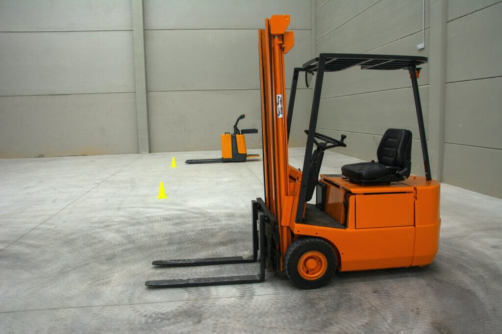 Een oranje heftruck staat binnen geparkeerd op een betonnen vloer, klaar voor drie situaties waarbij een heftruckchauffeur nodig is. Op de achtergrond zijn nog een oranje heftruck en twee gele verkeerskegels te zien. De grijze muren suggereren dat het een groot magazijn of opslagfaciliteit is.
