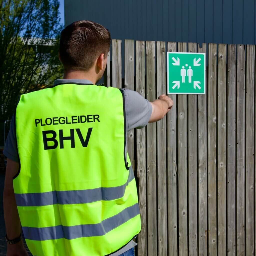 Een persoon in een felgeel veiligheidsvest met op de rug PLOEGLEIDER BHV wijst naar een bord met een noodverzamelpunt op een houten hek. Het tafereel weerspiegelt een van de 3 redenen om een BHV ploegleider te hebben: ervoor zorgen dat iedereen weet waar hij of zij heen moet in geval van nood.