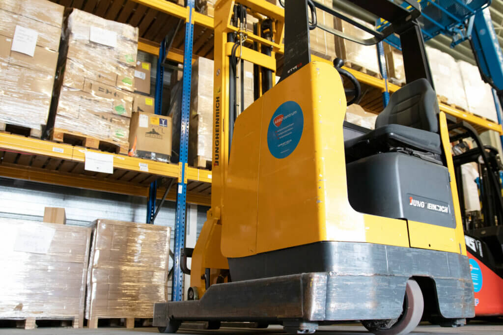 Een gele vorkheftruck staat geparkeerd in een magazijn, de omgeving is georganiseerd met planken vol dozen en pallets op blauwe metalen rekken. Het is een efficiënte setting die iemand ertoe kan aanzetten om 3 situaties te overwegen waarbij je een reachtruck gebruikt voor optimale materiaalbehandeling.