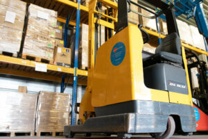 Een gele vorkheftruck staat geparkeerd in een magazijn, de omgeving is georganiseerd met planken vol dozen en pallets op blauwe metalen rekken. Het is een efficiënte setting die iemand ertoe kan aanzetten om 3 situaties te overwegen waarbij je een reachtruck gebruikt voor optimale materiaalbehandeling.